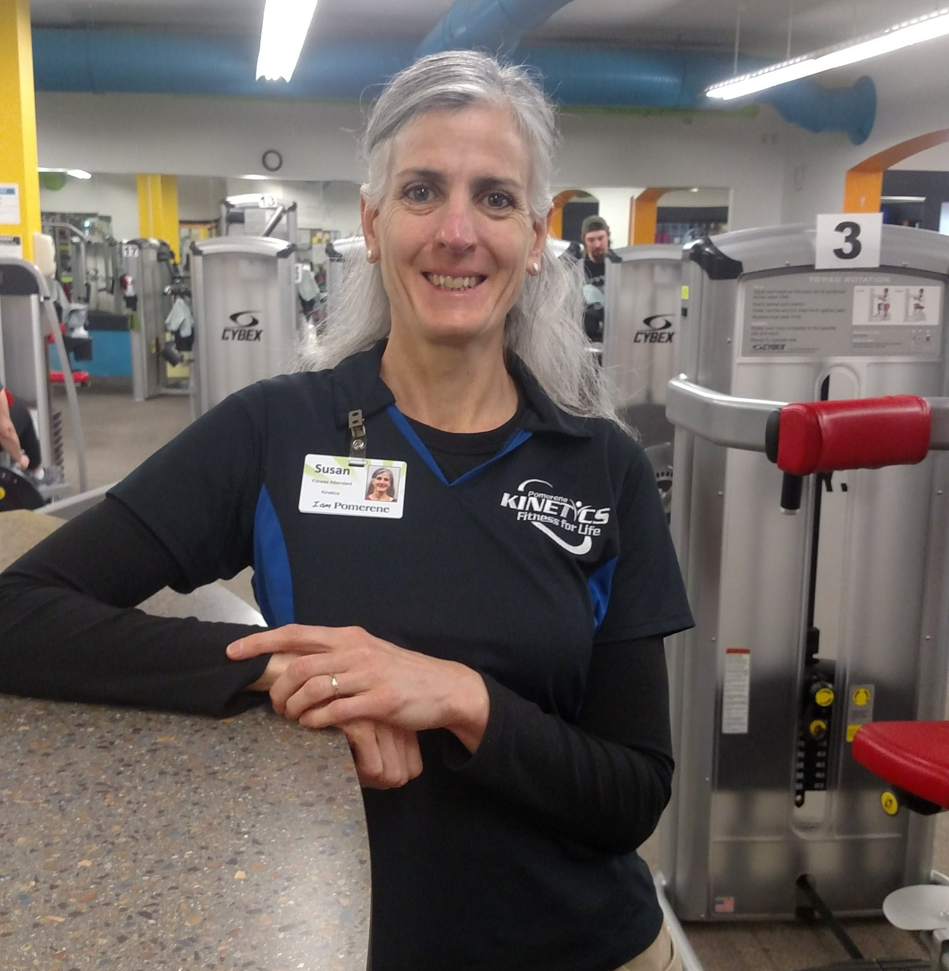 Woman in gym uniform, smiling, leans on counter near exercise equipment.  She is wearing a nametag and is in a gym setting.