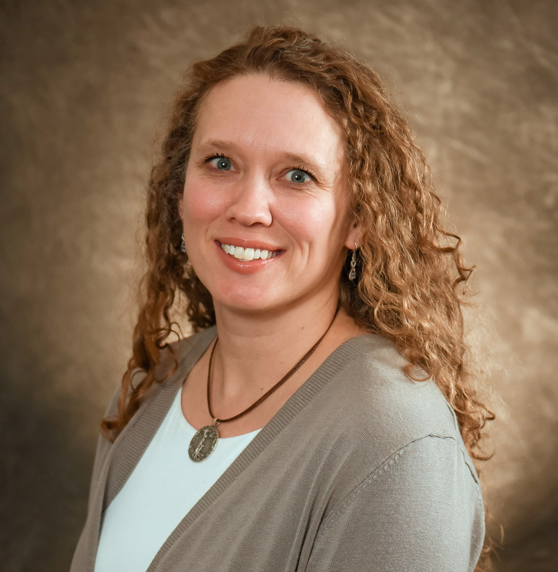 Woman with curly brown hair smiles, wearing a white shirt and gray cardigan, against a brown backdrop.
