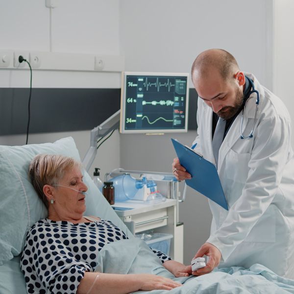 Young woman in a white coat with a stethoscope, holding a clipboard, smiling at the camera in a hospital setting.