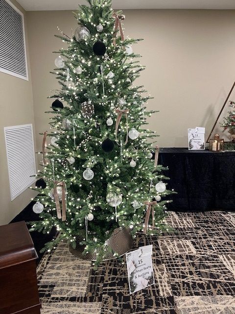 Christmas tree decorated with lights, black, silver, and tan ornaments, indoors. A sign sits at the base of the tree.
