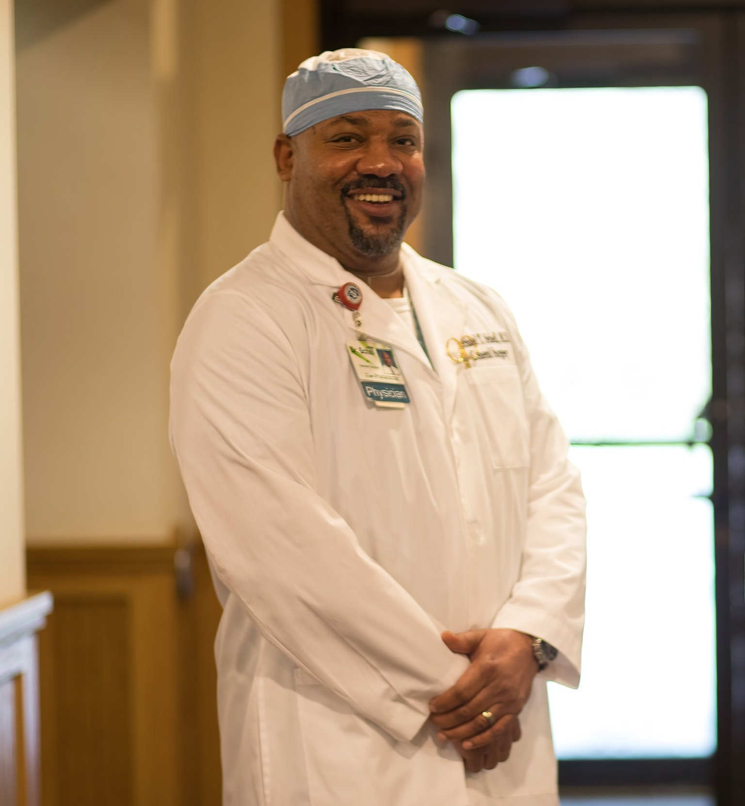 Smiling Black doctor wearing a surgical cap and white coat, standing in a hallway.
