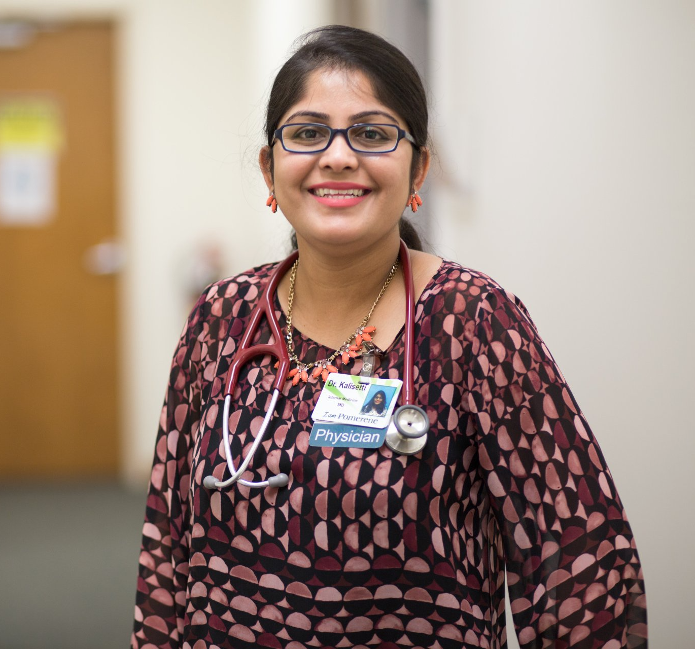 Woman doctor in glasses, wearing stethoscope and a patterned top, smiling in a hospital hallway.
