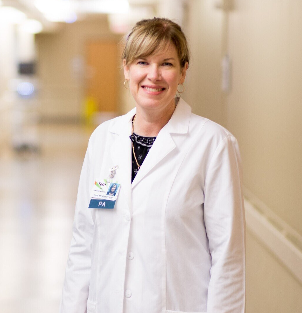 A woman in a white coat, a PA badge, smiles in a hospital hallway. She has blonde hair and wears a patterned blouse.