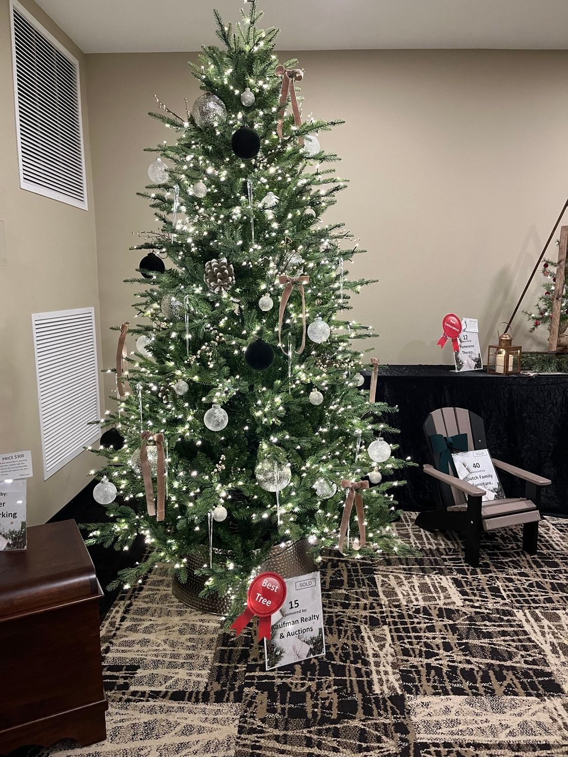 Christmas tree decorated with lights and ornaments, in a room with art and a small wooden chair.