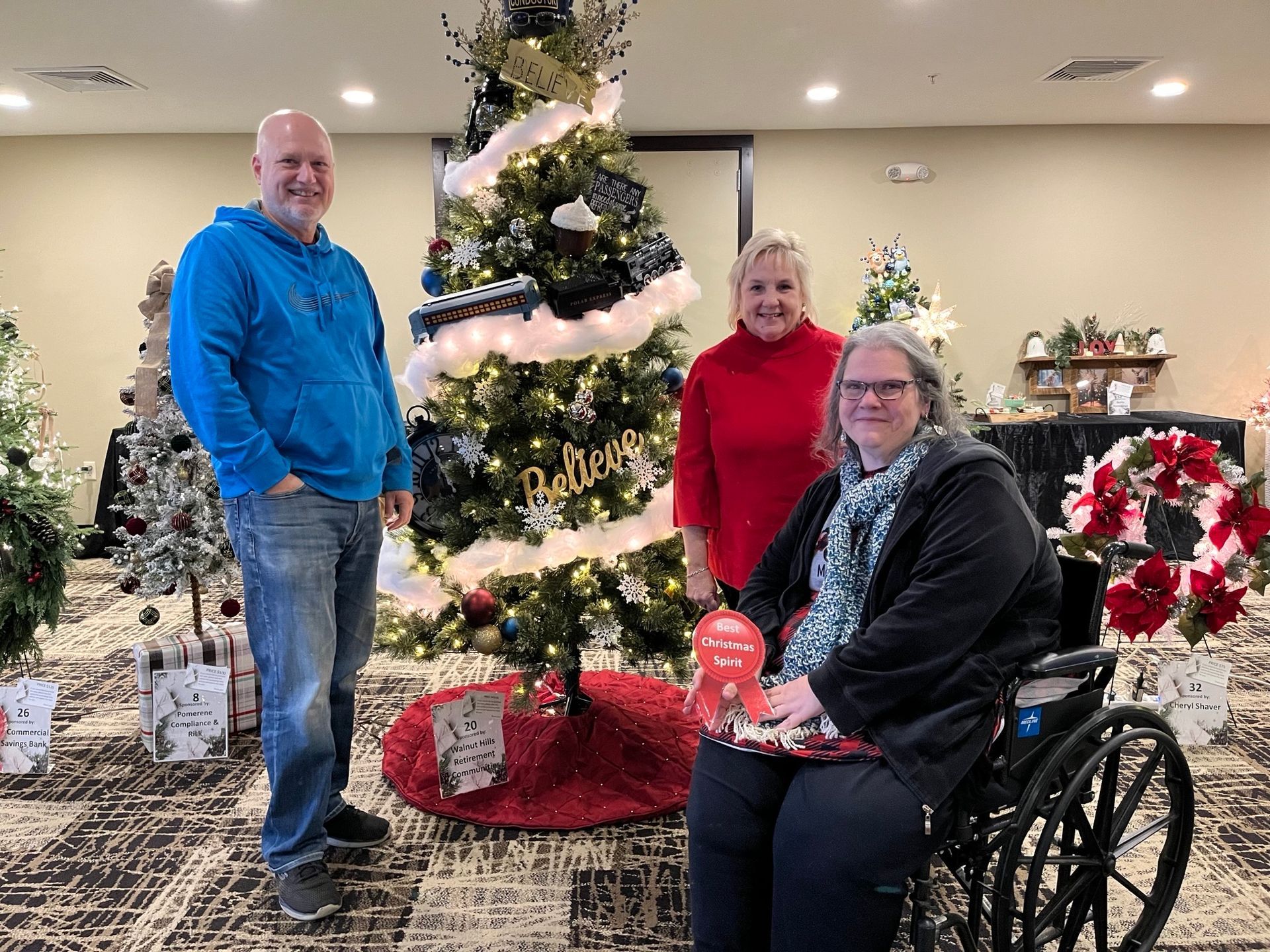 Three people pose by a Christmas tree. A woman in a wheelchair holds a gift. Festive setting.