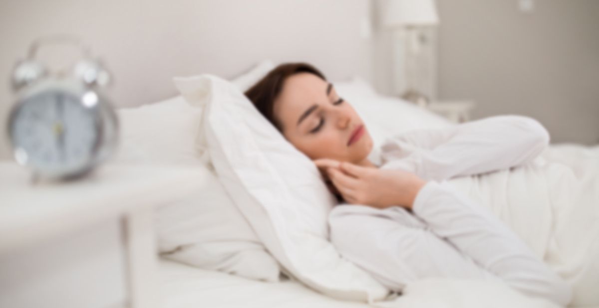Woman sleeping in bed next to an alarm clock, wearing white clothing in a bright room.