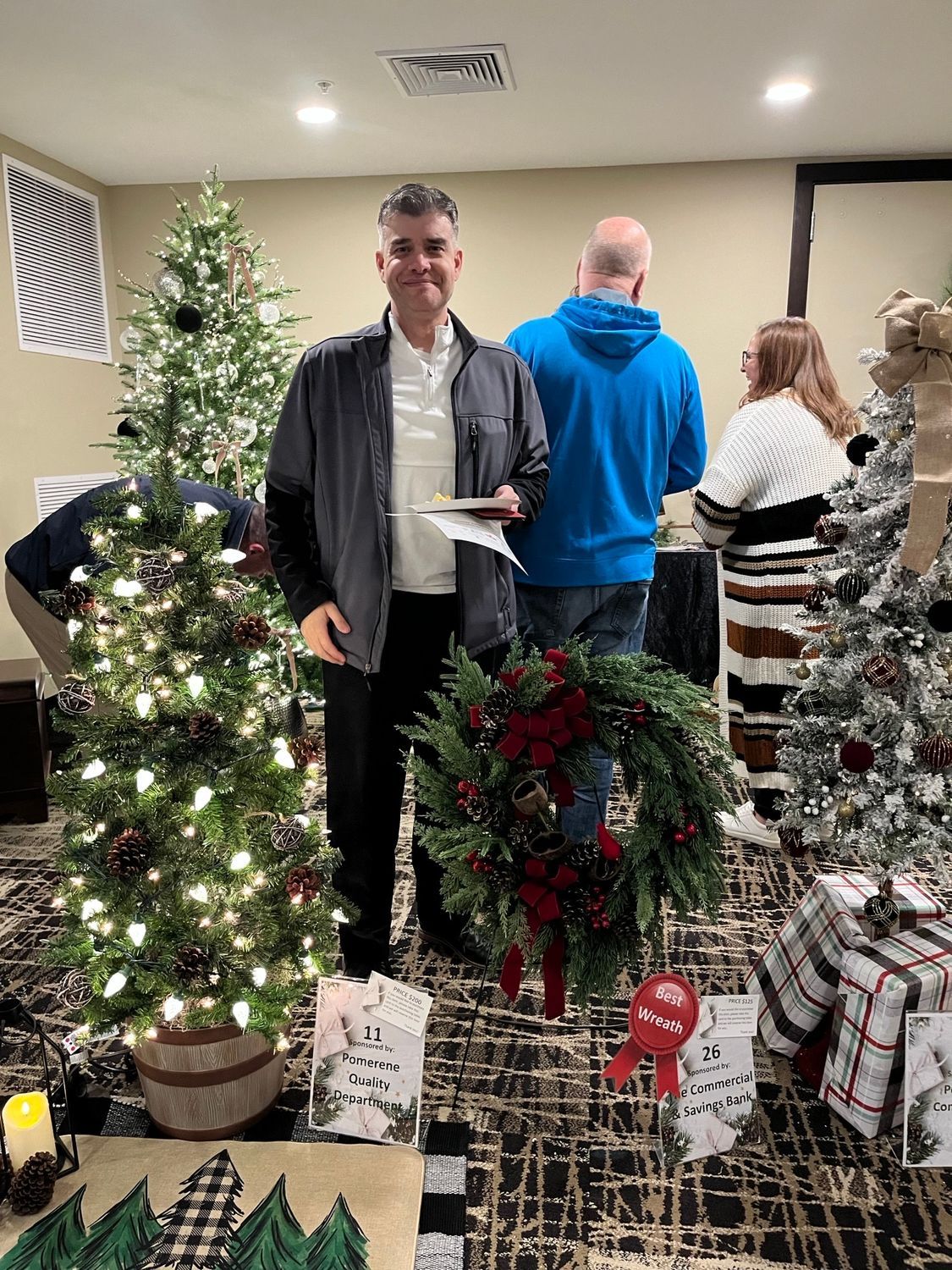 A man in a denim jacket smiles, holding a paper, standing near a Christmas tree and wreath. Others are in the decorated room.