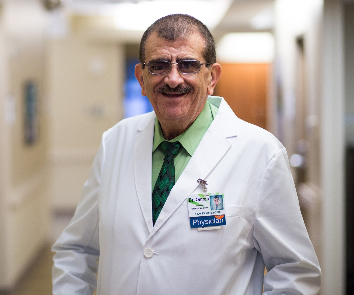 Man in white lab coat smiling in a hospital hallway, wearing glasses and a green tie.