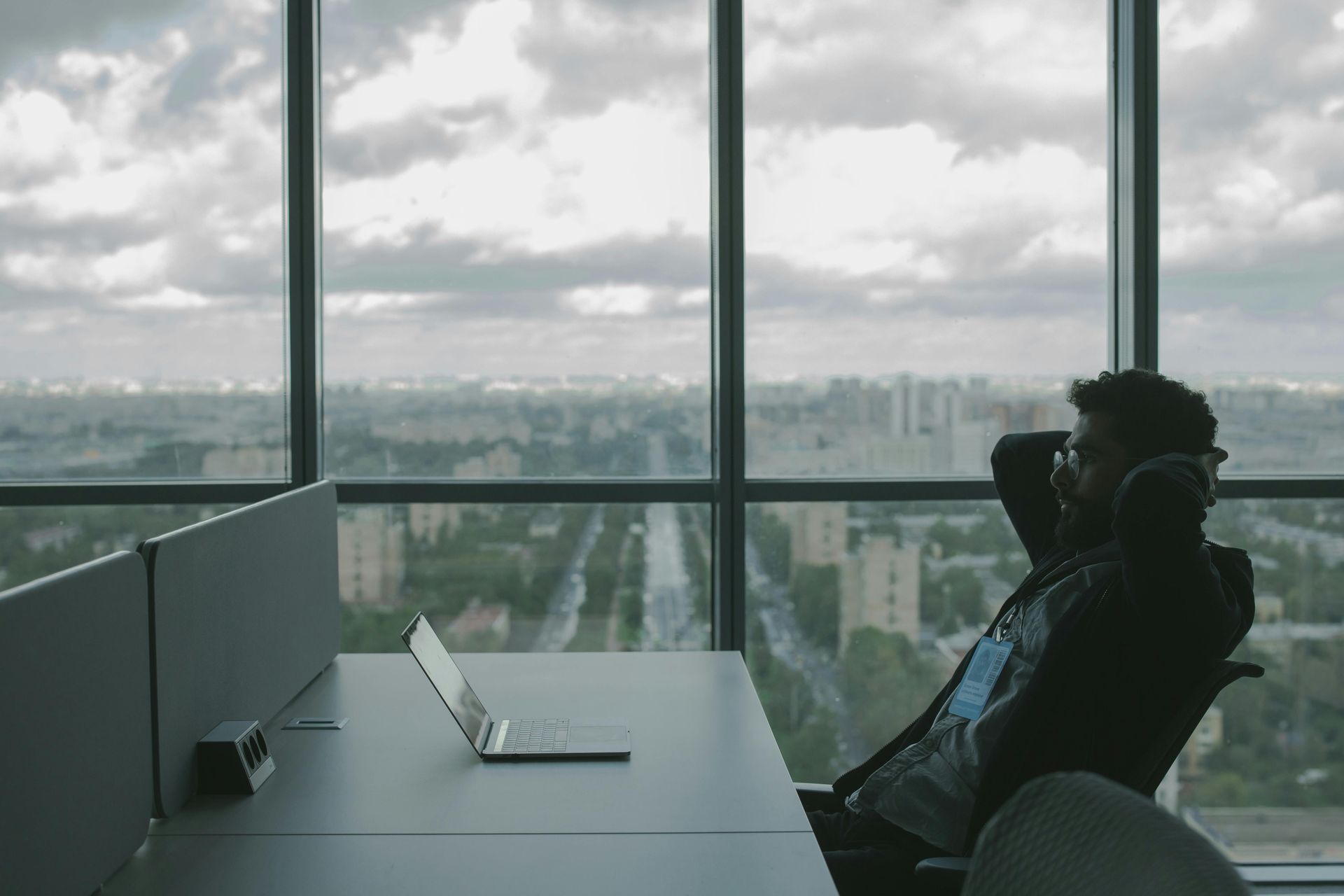 Man relaxing in office chair, looking out large window at cityscape with laptop on desk.