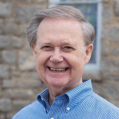 A man in a blue and white striped shirt is smiling in front of a stone wall.