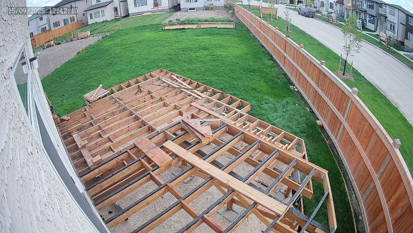 Deck frame under construction in a backyard, surrounded by grass, a fence, and houses.