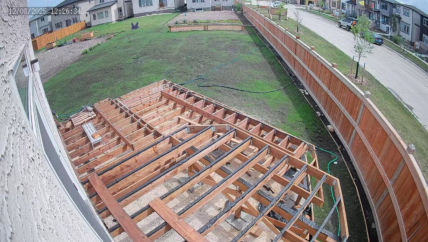 Overhead view of a deck frame under construction, surrounded by a lawn, fence, and buildings. Brown wood and green grass.