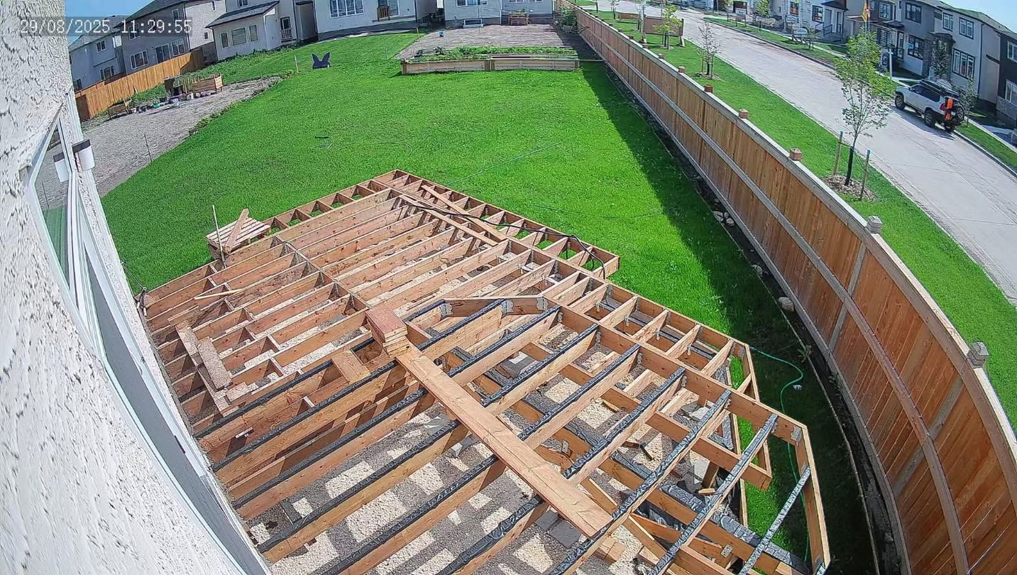 A partially built house frame with green lawn, wooden fence, and nearby houses.
