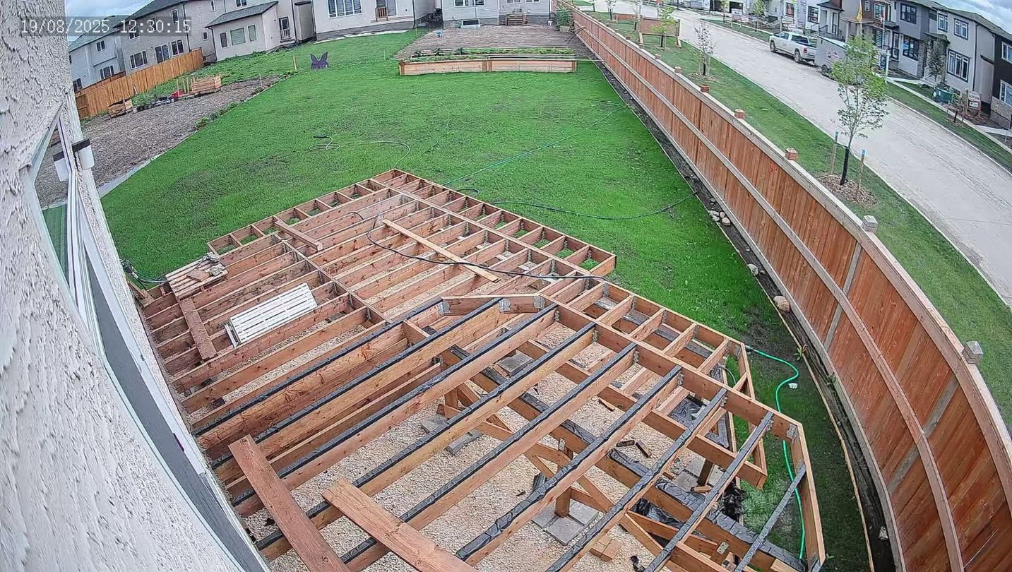 Framed wooden deck construction in a backyard with green grass, bordered by a brown fence and buildings.