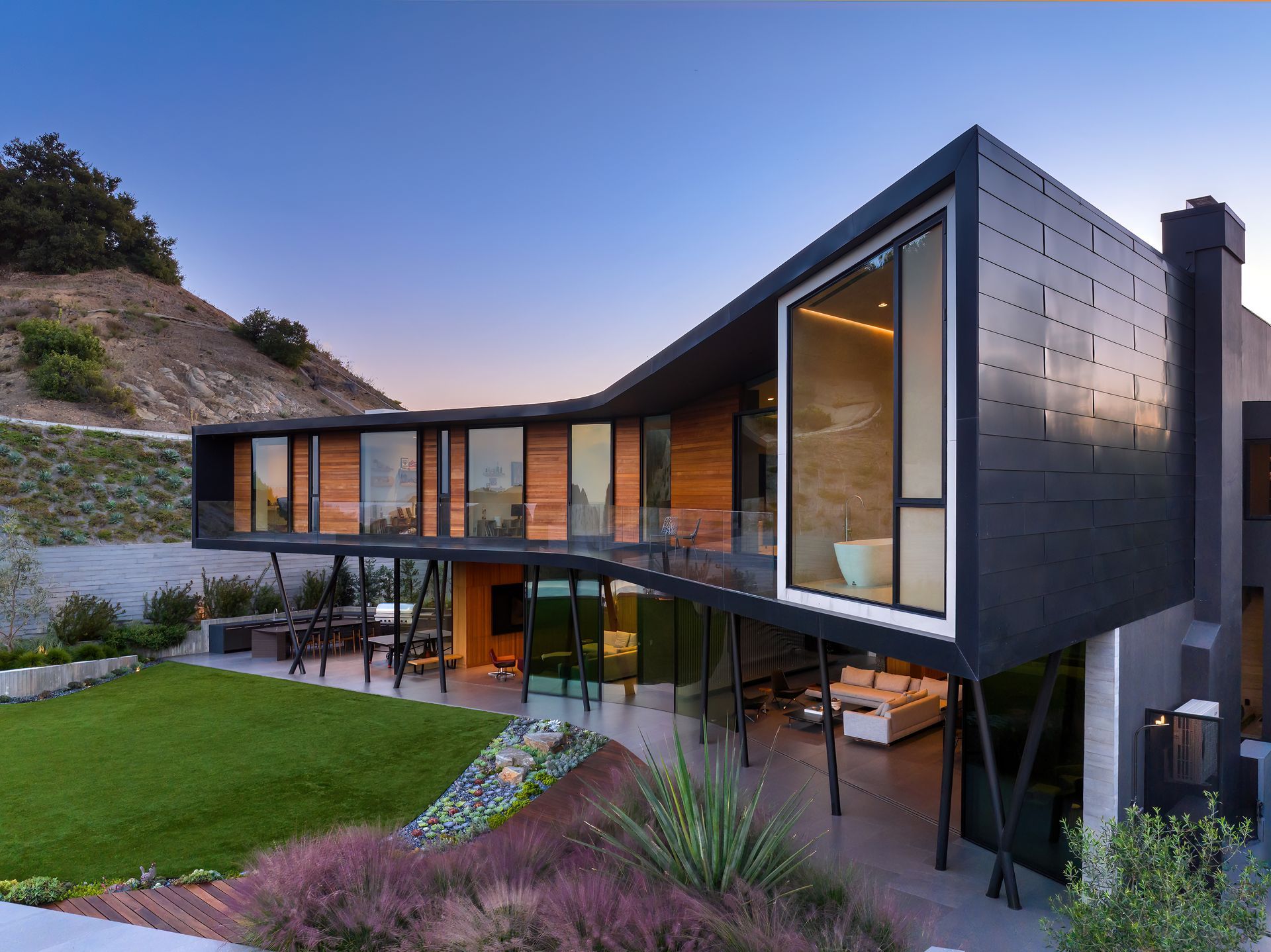 Modern hillside house with large glass walls, courtyard lawn, and warm interior lights at dusk