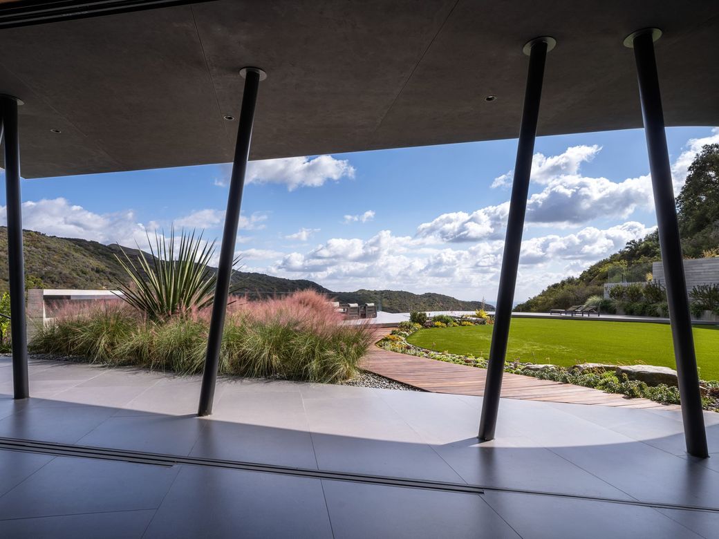 Modern patio with black columns overlooking landscaped garden, pink flowers, and a cloudy blue sky