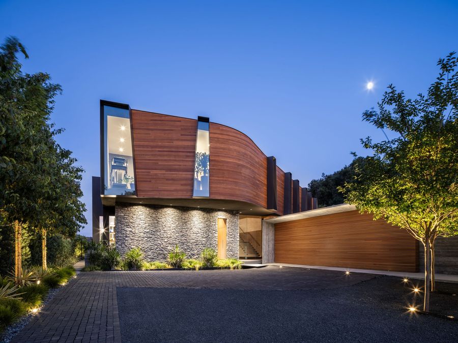 Modern stone-and-wood house at dusk with warm exterior lights and a curved driveway