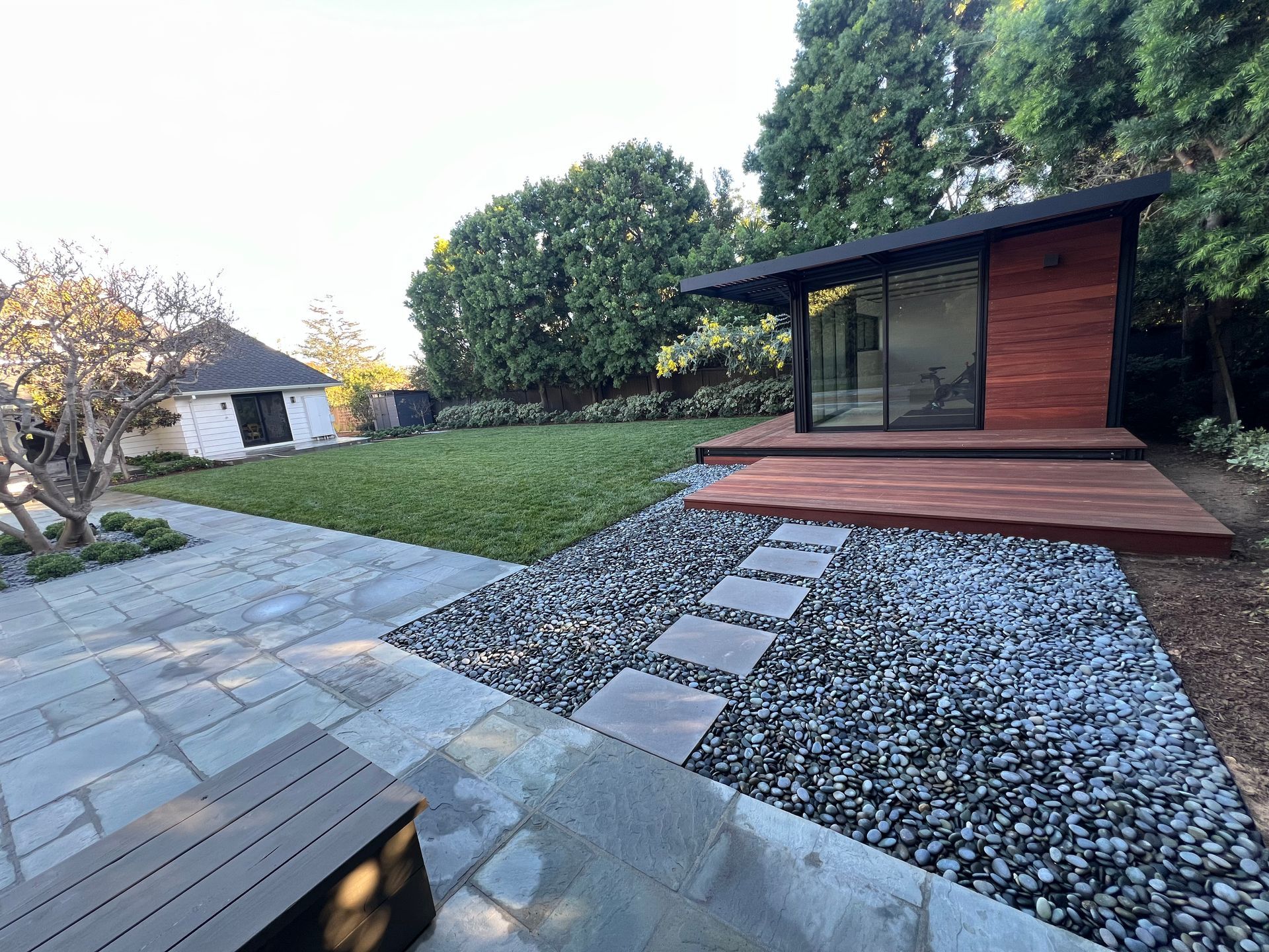 Modern backyard with gray stone path, gravel bed, and small wood deck beside a sleek garden shed.