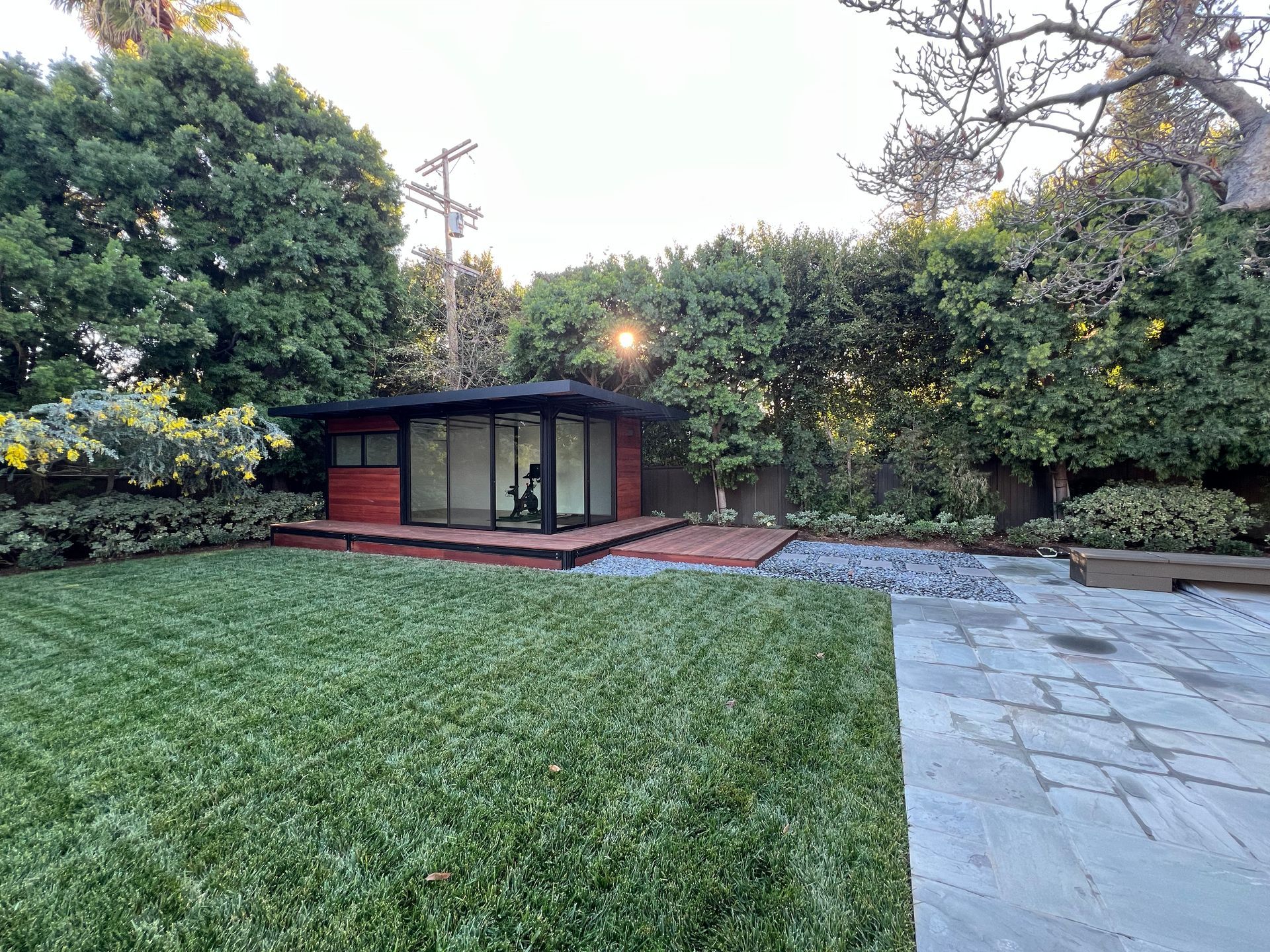 Backyard with a small red garden shed, green lawn, hedges, and stone patio at dusk