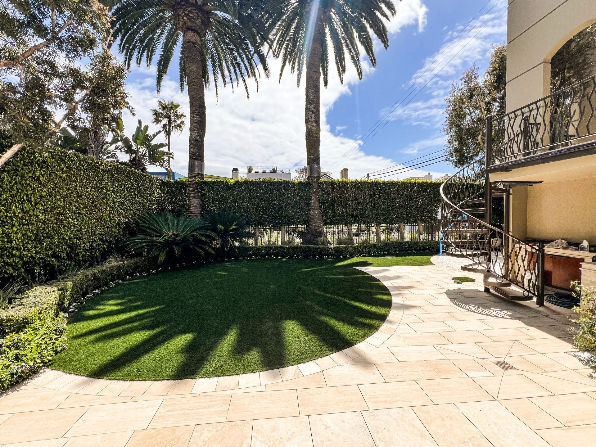 Sunny patio with palm trees, circular lawn, hedge, and curved staircase beside a beige house