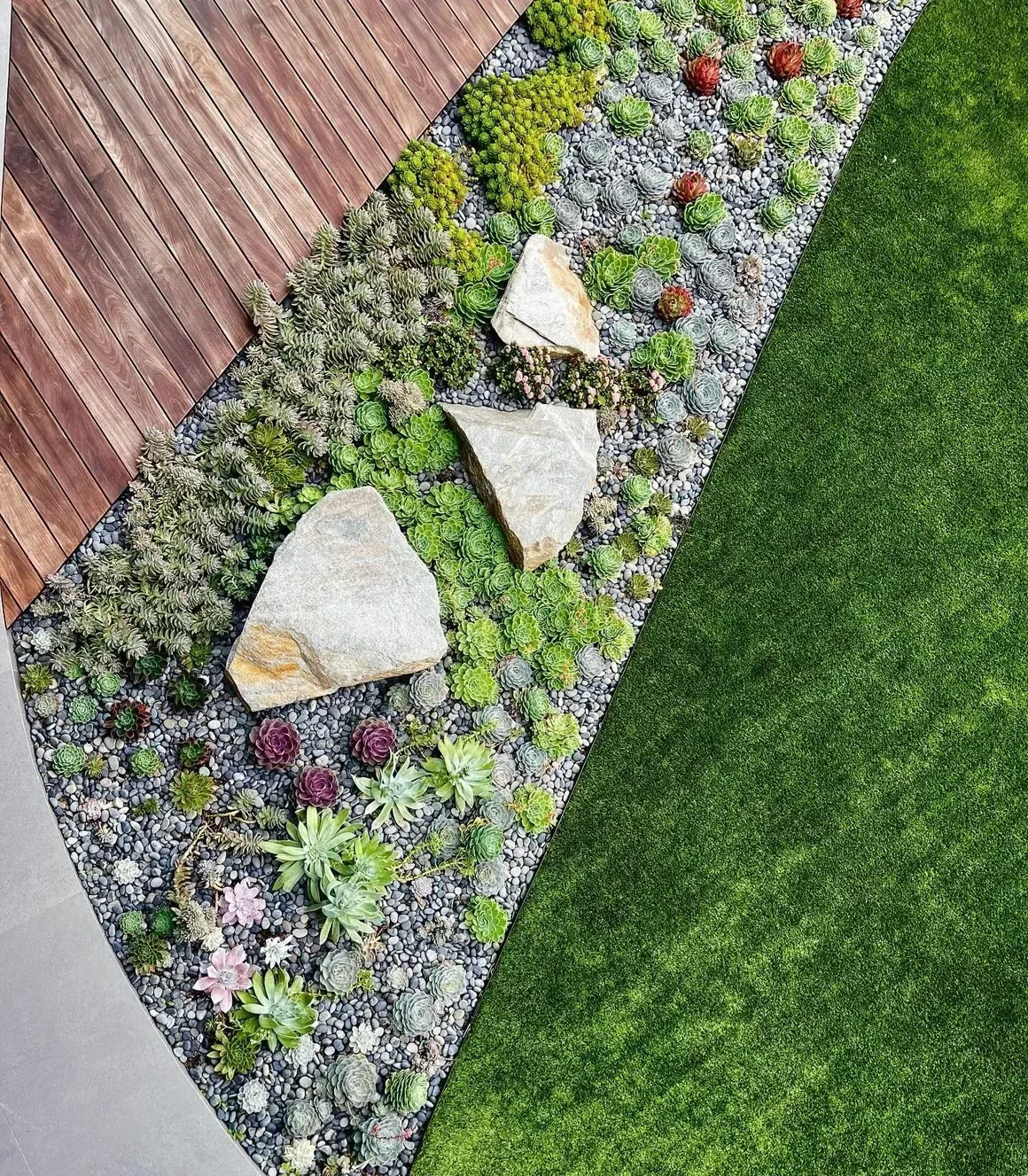 Landscaped garden border with stones, colorful flowers, and a wooden deck beside green lawn
