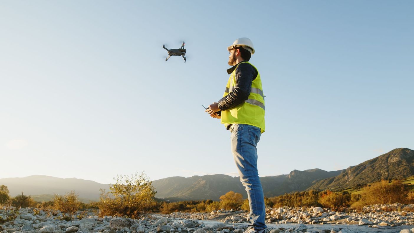 A person in a hard hat and high-visibility vest standing in a rocky landscape while flying a drone.