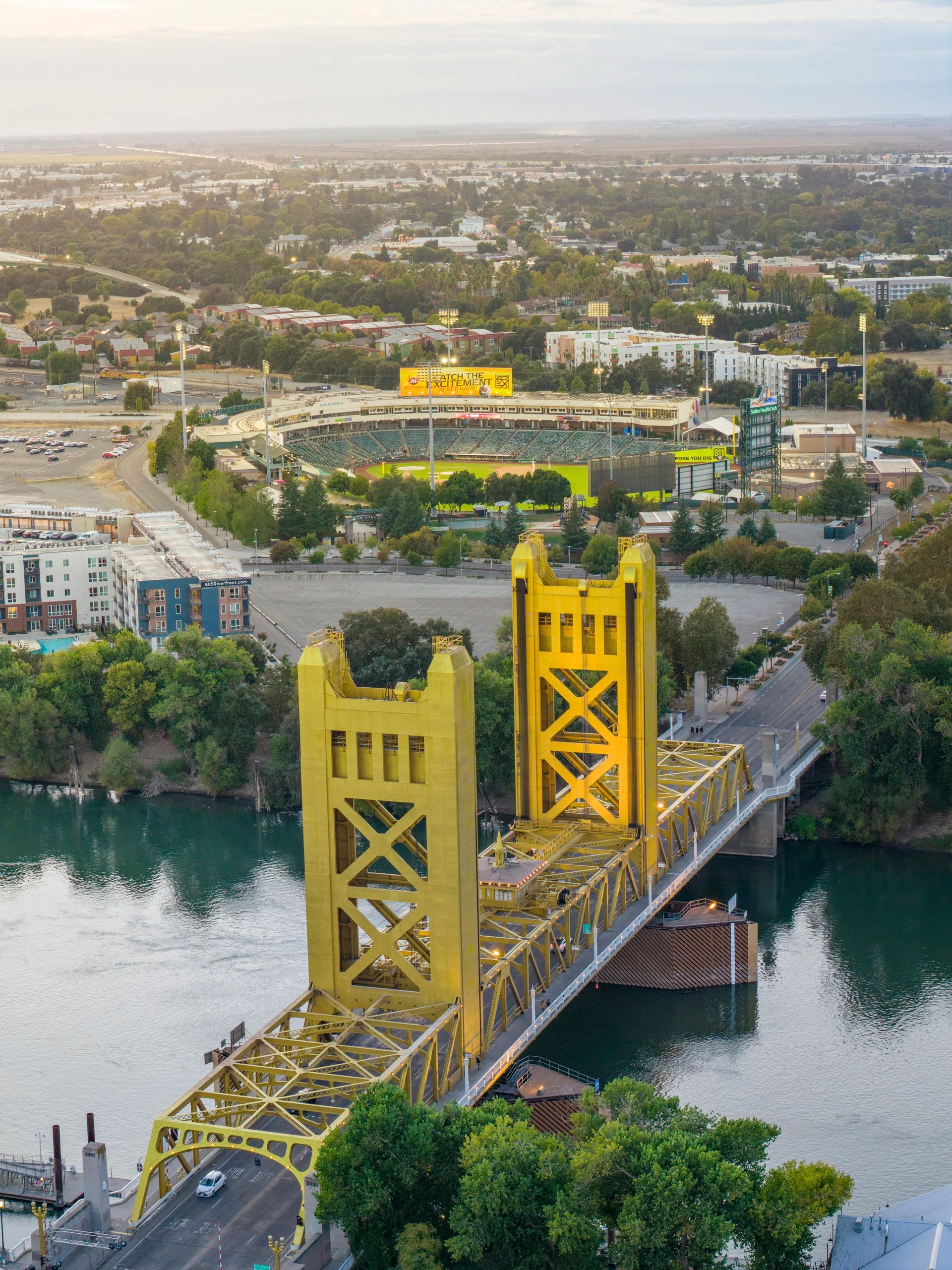 An aerial view of the bright yellow Tower Bridge over the Sacramento River, with a stadium visible in the background.