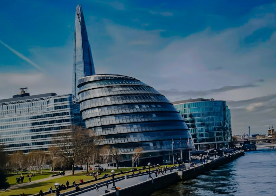 London cityscape: The Shard skyscraper rises behind curved City Hall, both on the Thames waterfront under a blue sky.