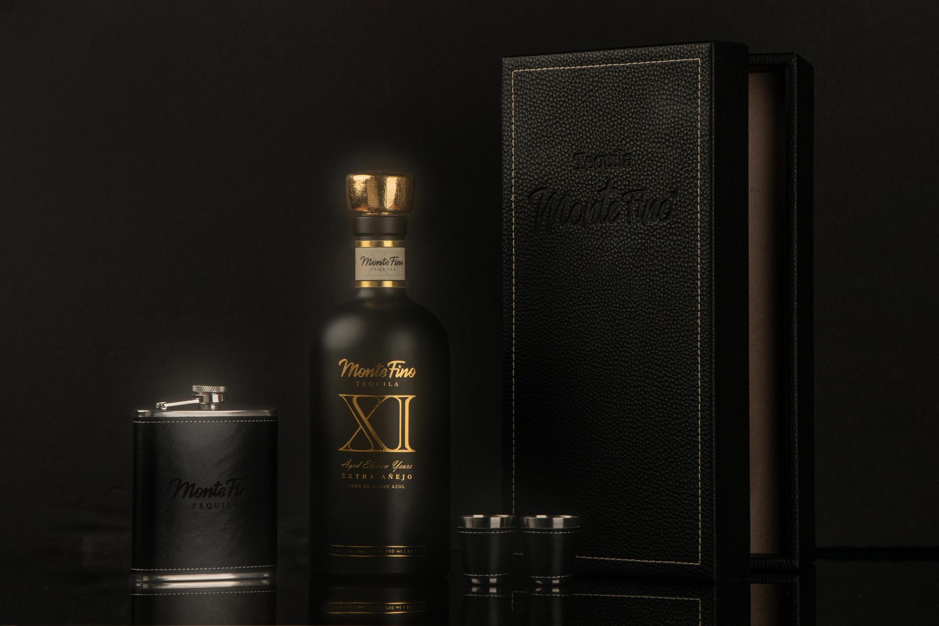 Black liquor bottle, flask, and glasses with gold accents on a dark surface.