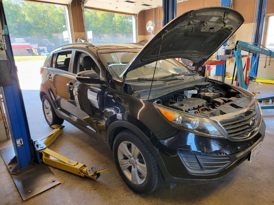 Black Kia SUV with open hood, in a repair shop, raised on a lift.