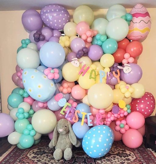 A teddy bear is sitting in front of a wall of balloons that say happy easter