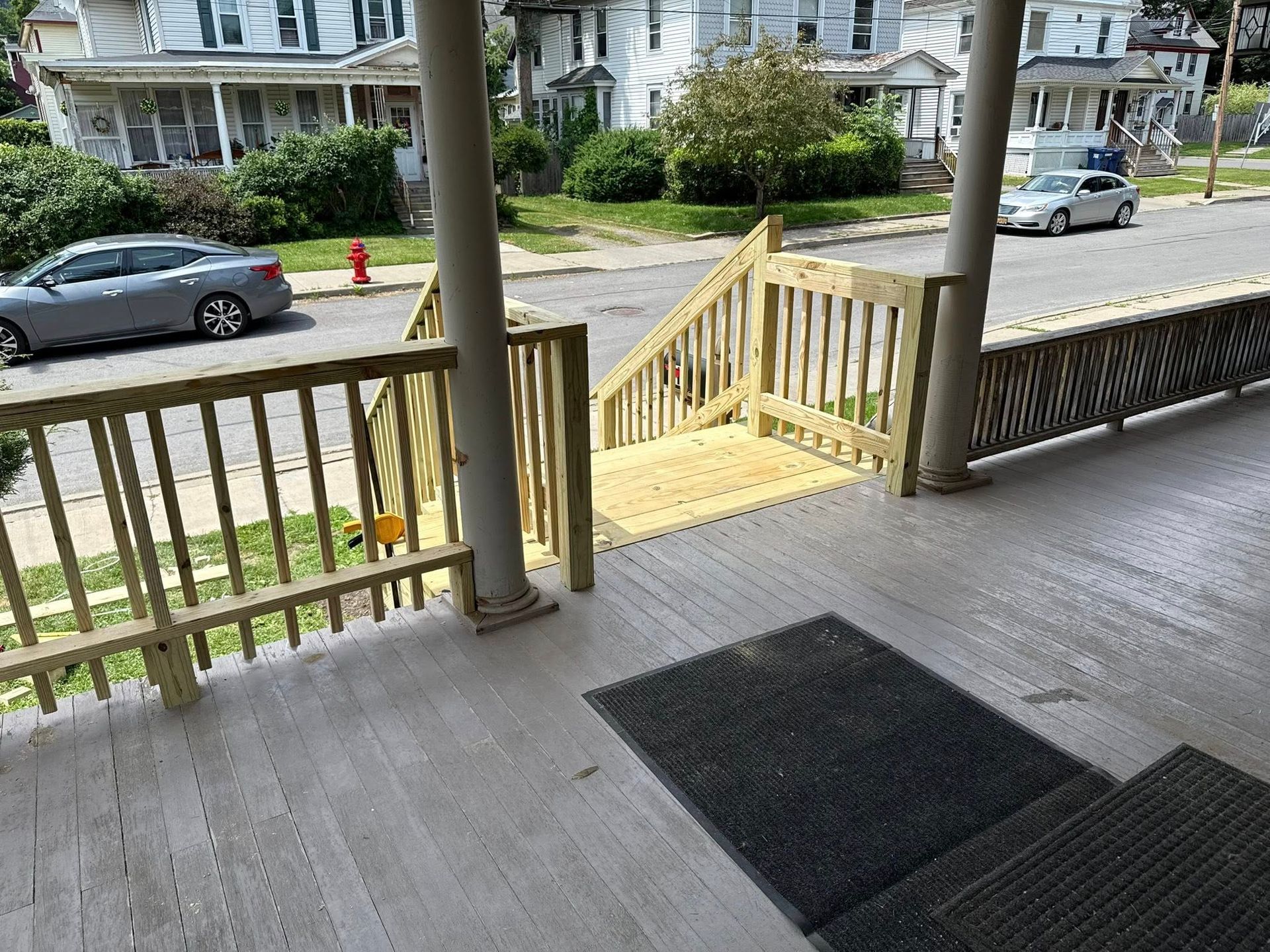Wooden porch with stairs, railing, and a doormat. Buildings and cars are visible in the background.