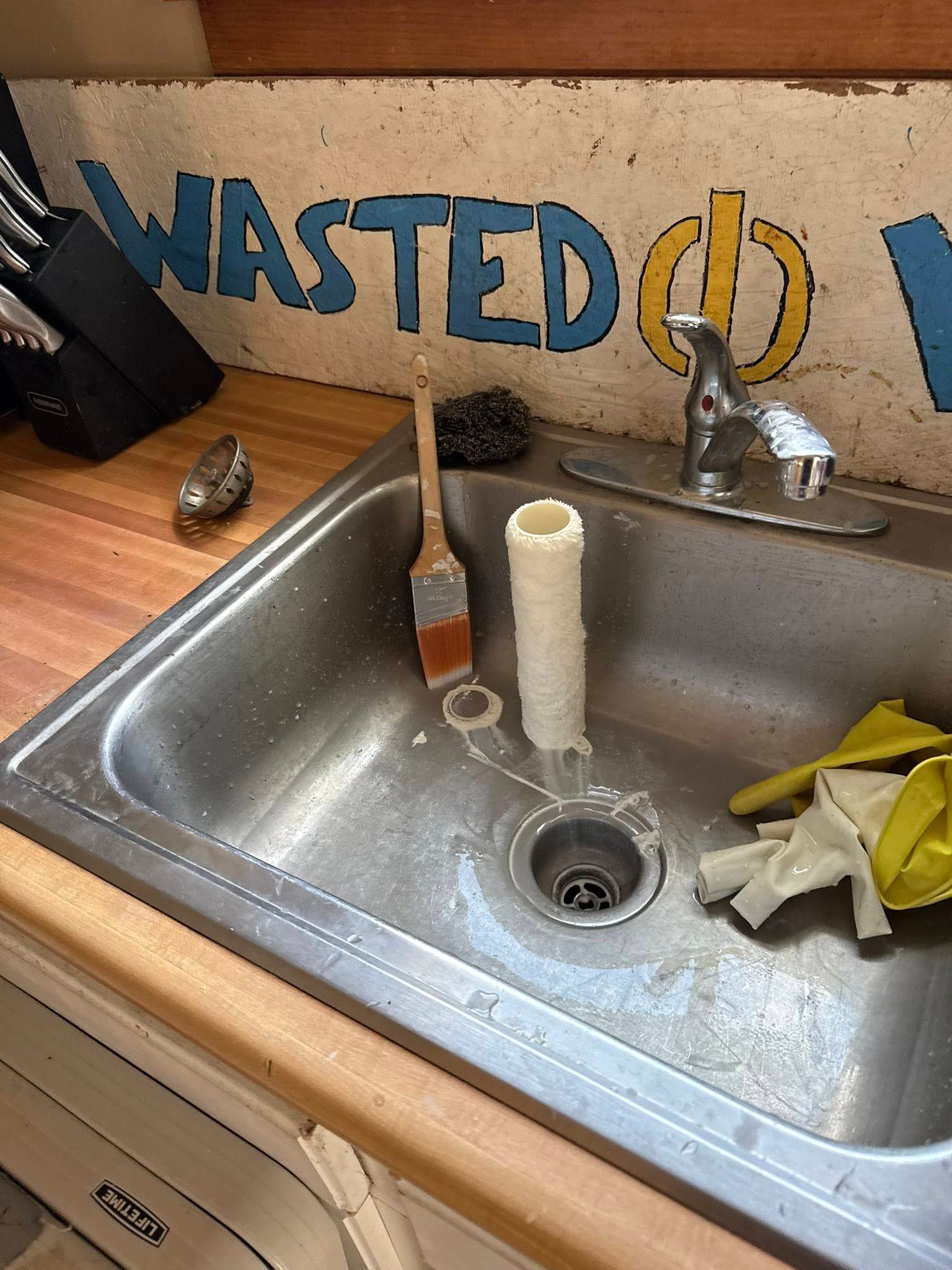 A cluttered stainless steel sink with a paint brush, sponge, and a partial PVC pipe.