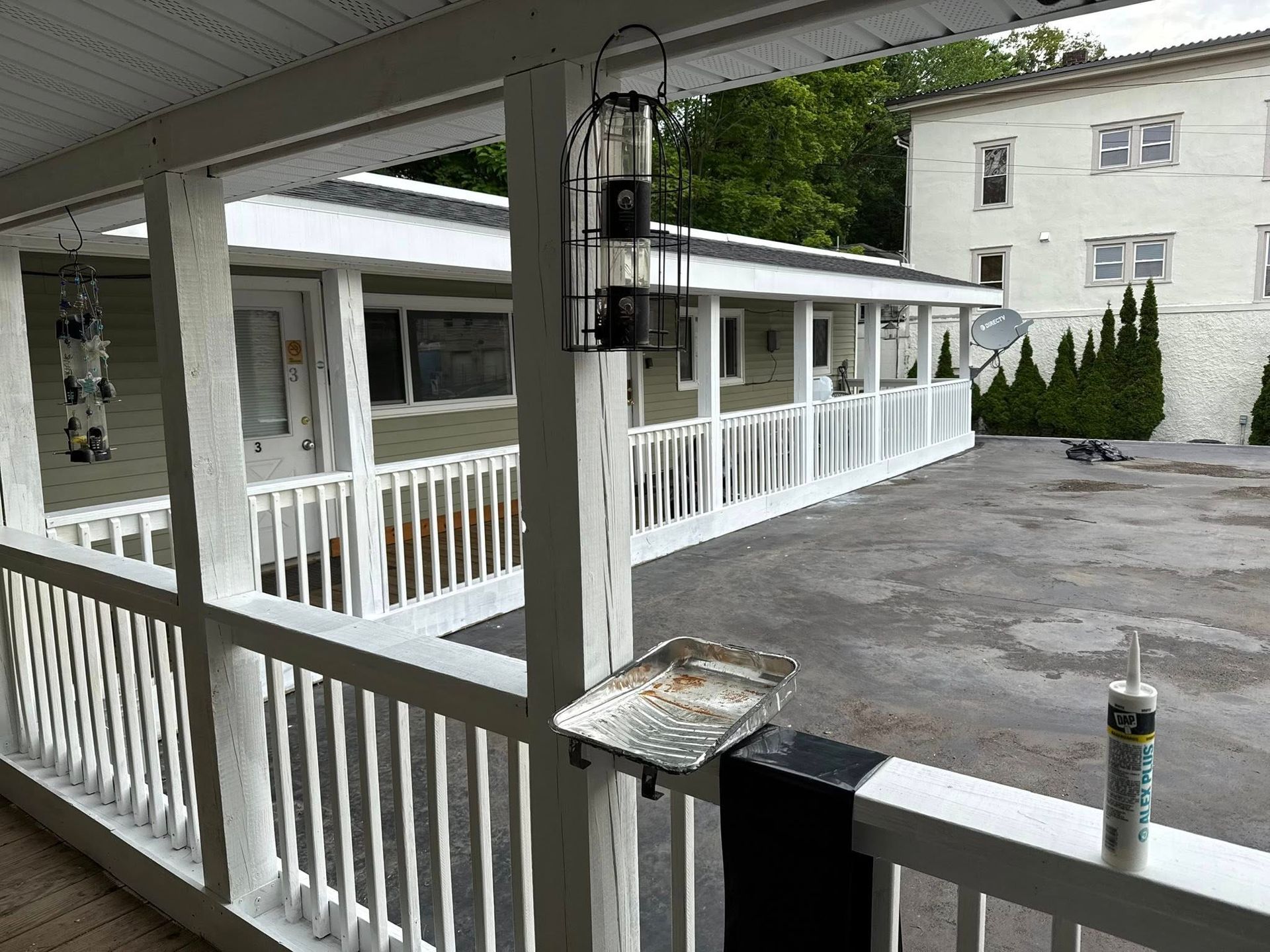 A porch with white railing overlooks a concrete area and a single-story building. Black bird feeder hangs.
