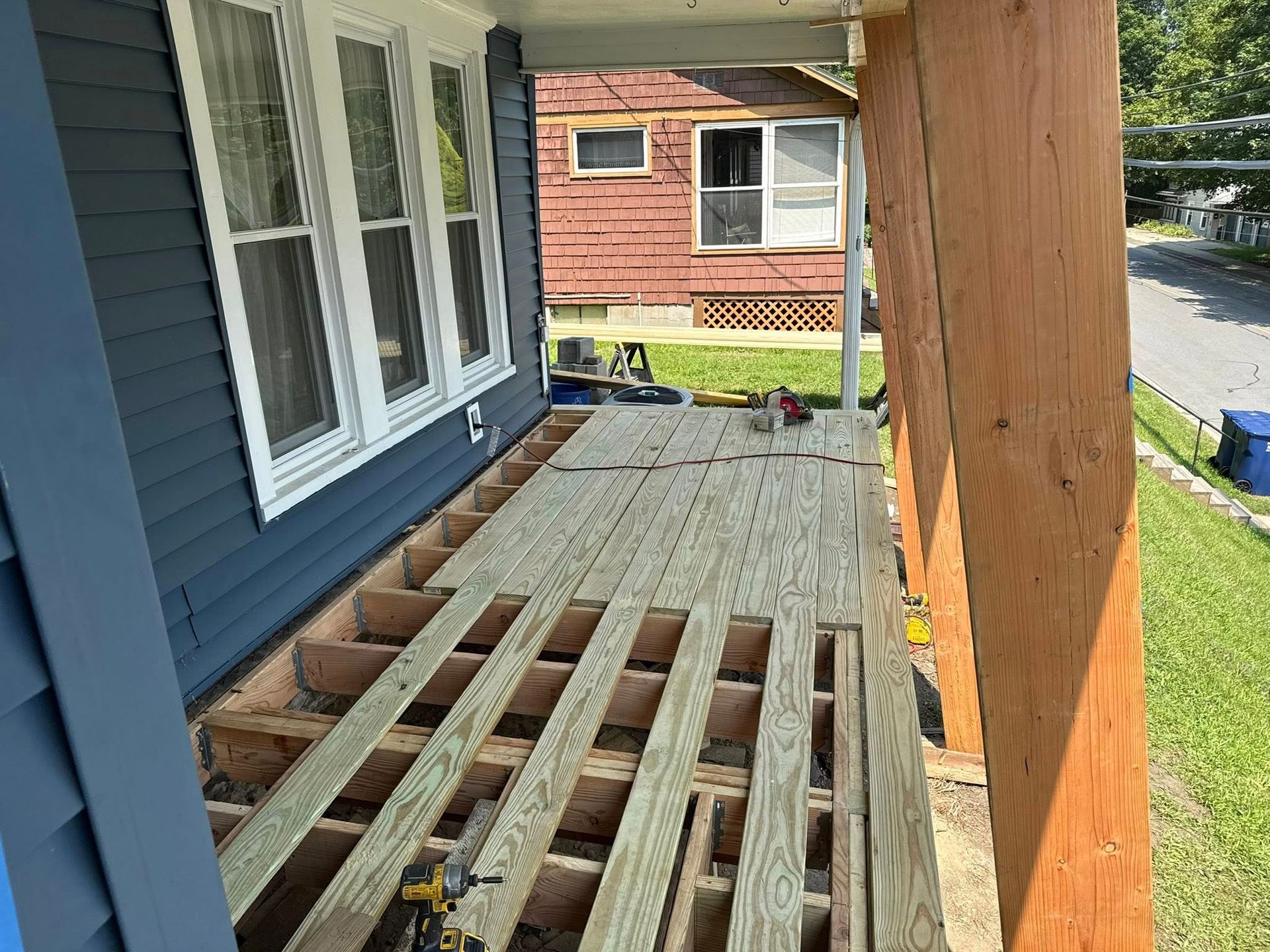 Porch under construction; exposed wood framing and deck. Blue house, brown post, and yard in background.