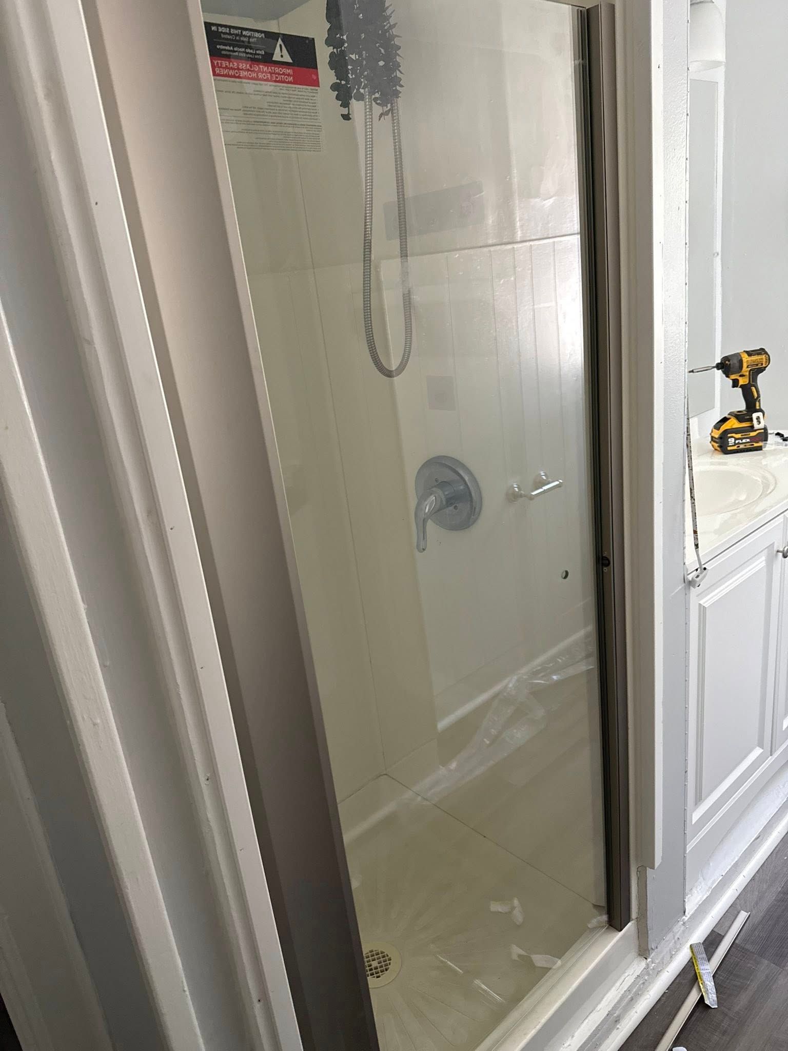 Shower door being installed. White frame, clear glass, drill visible.