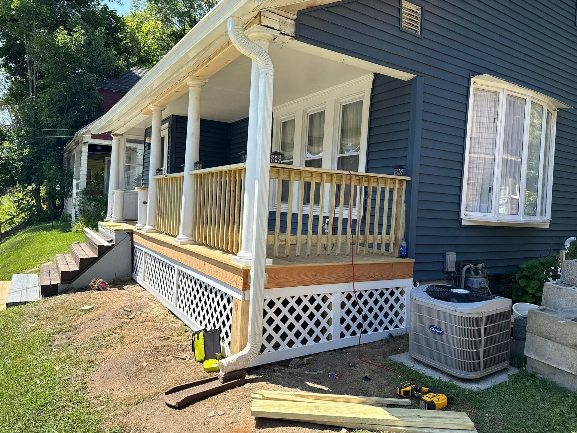 House with porch under construction, featuring new wooden railing and latticework, with HVAC unit nearby.