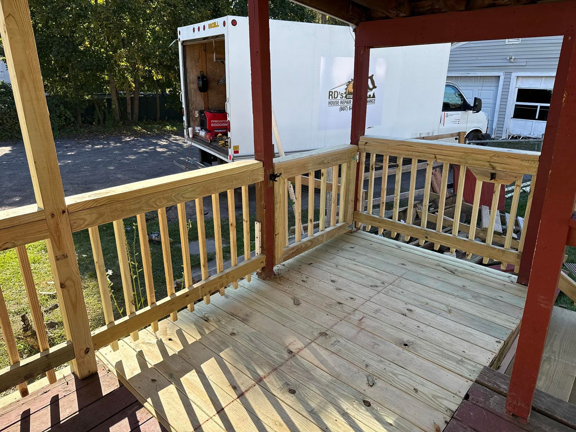 Newly constructed wooden porch with railing, adjacent to a white utility trailer.