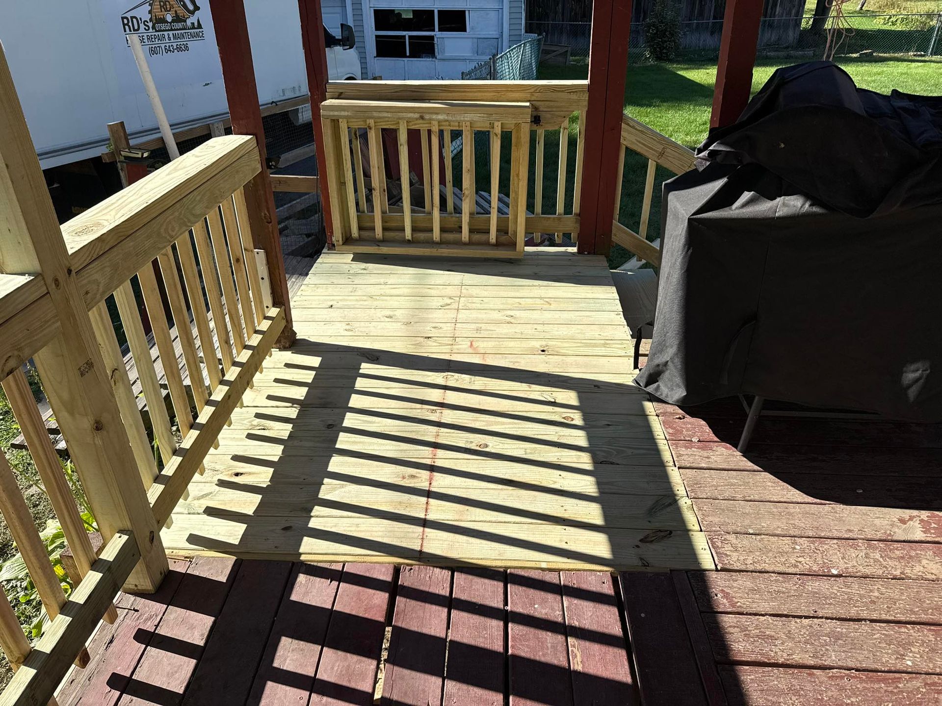 Wooden deck with railings, shaded by a porch roof, and a barbeque grill covered.
