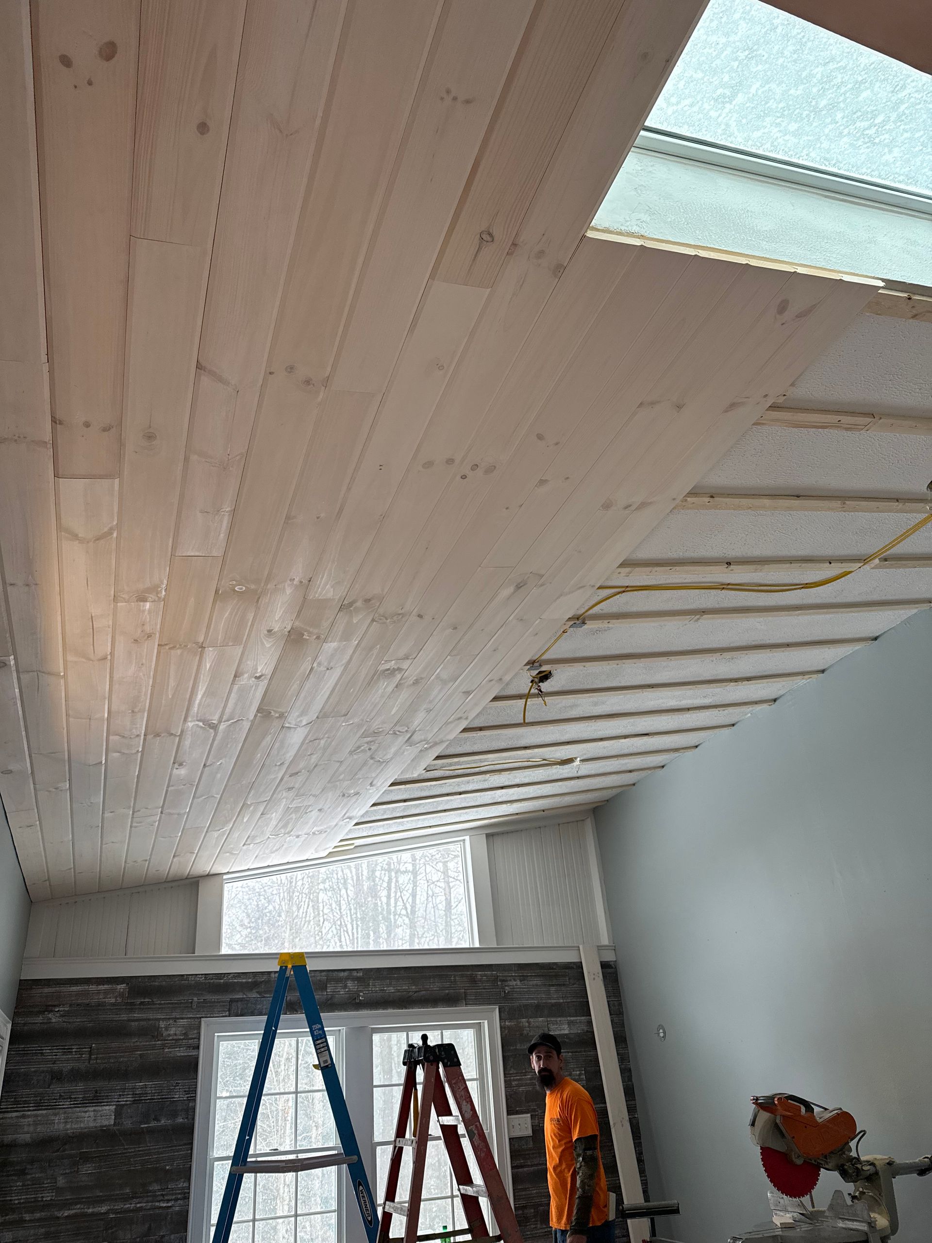 Ceiling wood paneling installation in progress, worker on ladder, skylight, interior setting.
