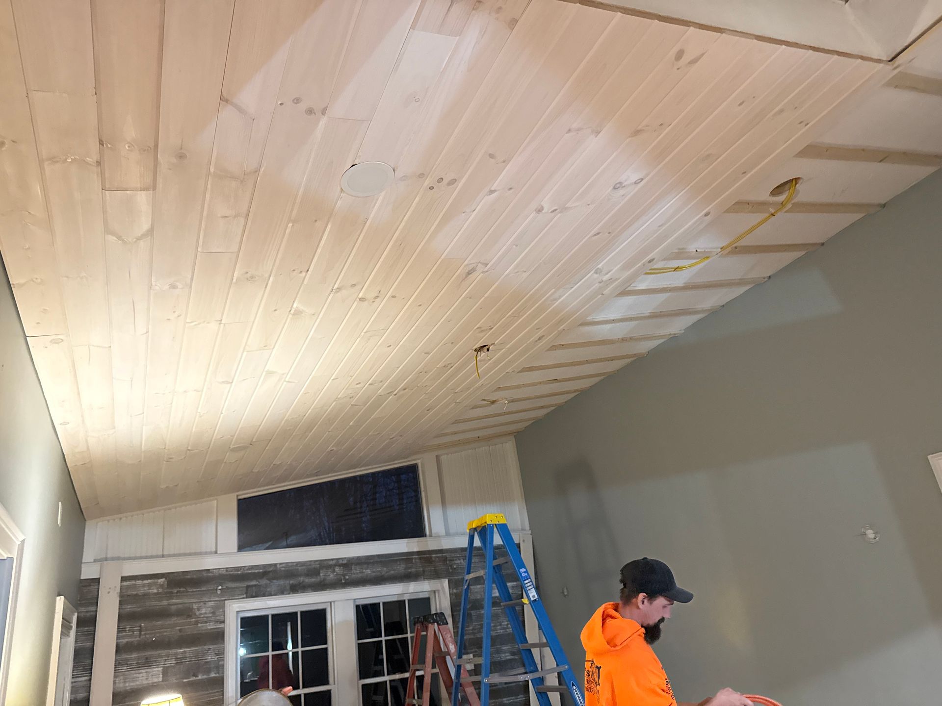 Man installing wooden planks on a sloped ceiling. Interior room with ladder and completed wood paneling.