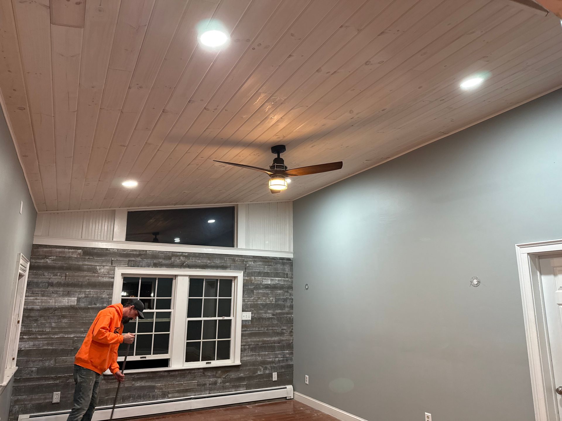 Man in orange shirt works on a room with wood ceiling, gray walls, and a window.