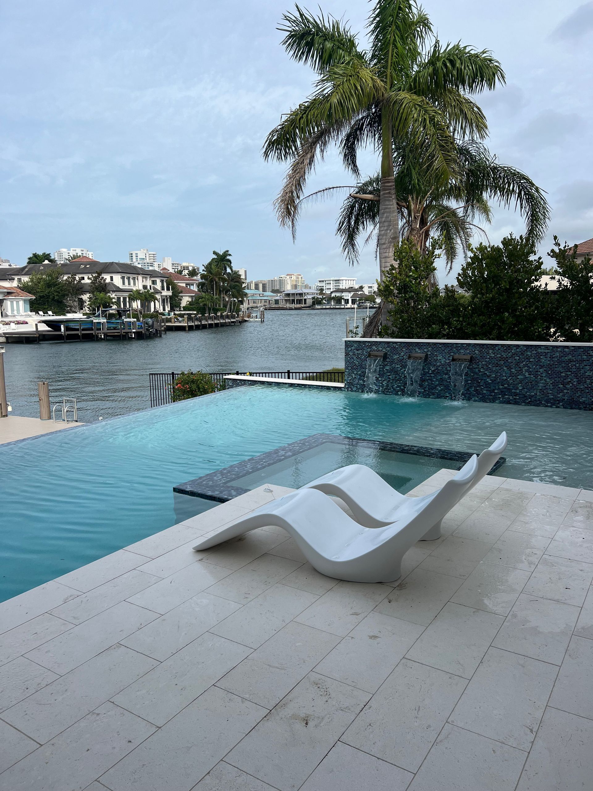 Poolside view: White chaise lounges, blue water, palm tree, waterway, buildings in the distance.