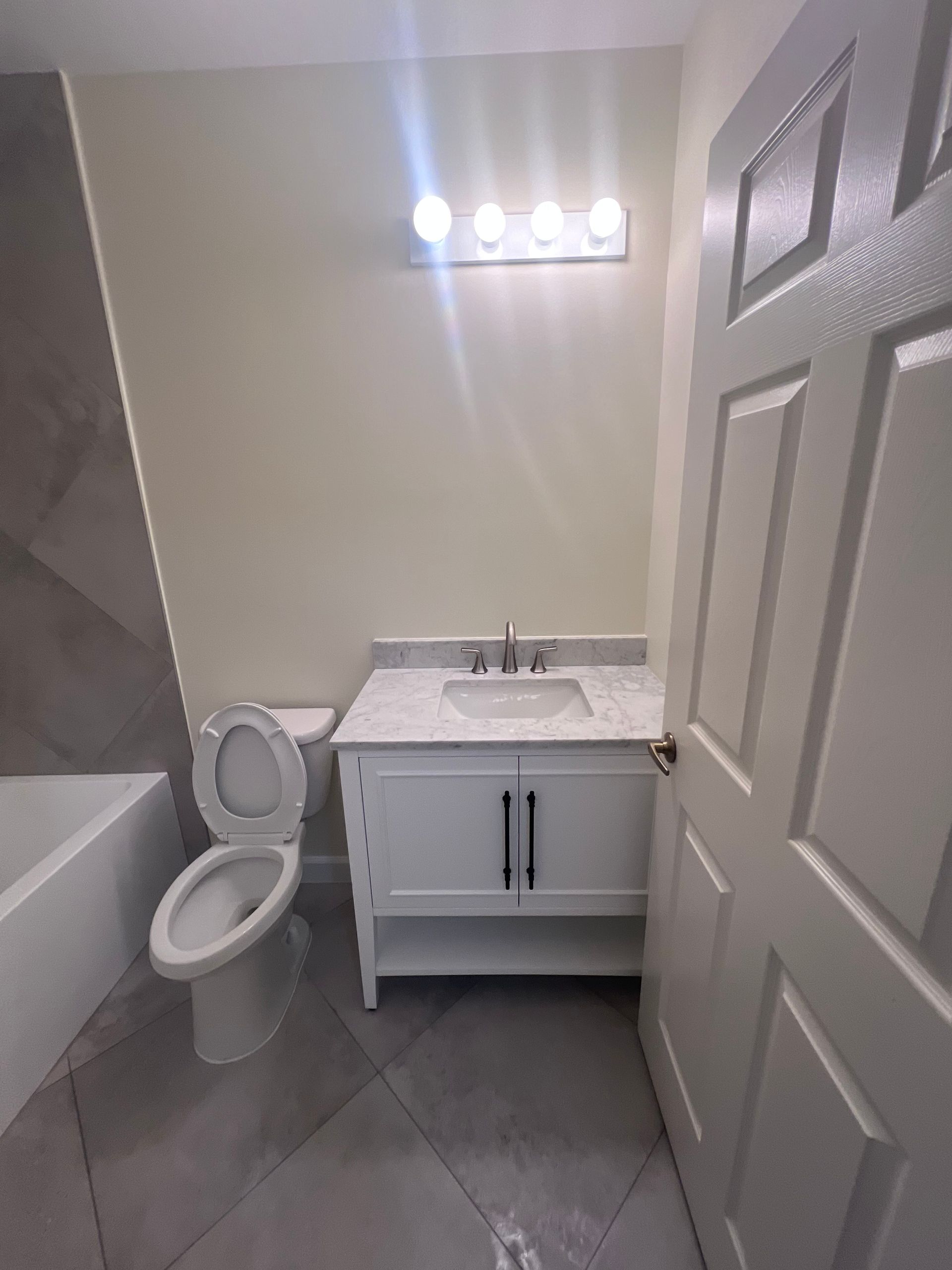 Bathroom with white vanity, toilet, and bathtub. Off-white walls, gray tiled floor.