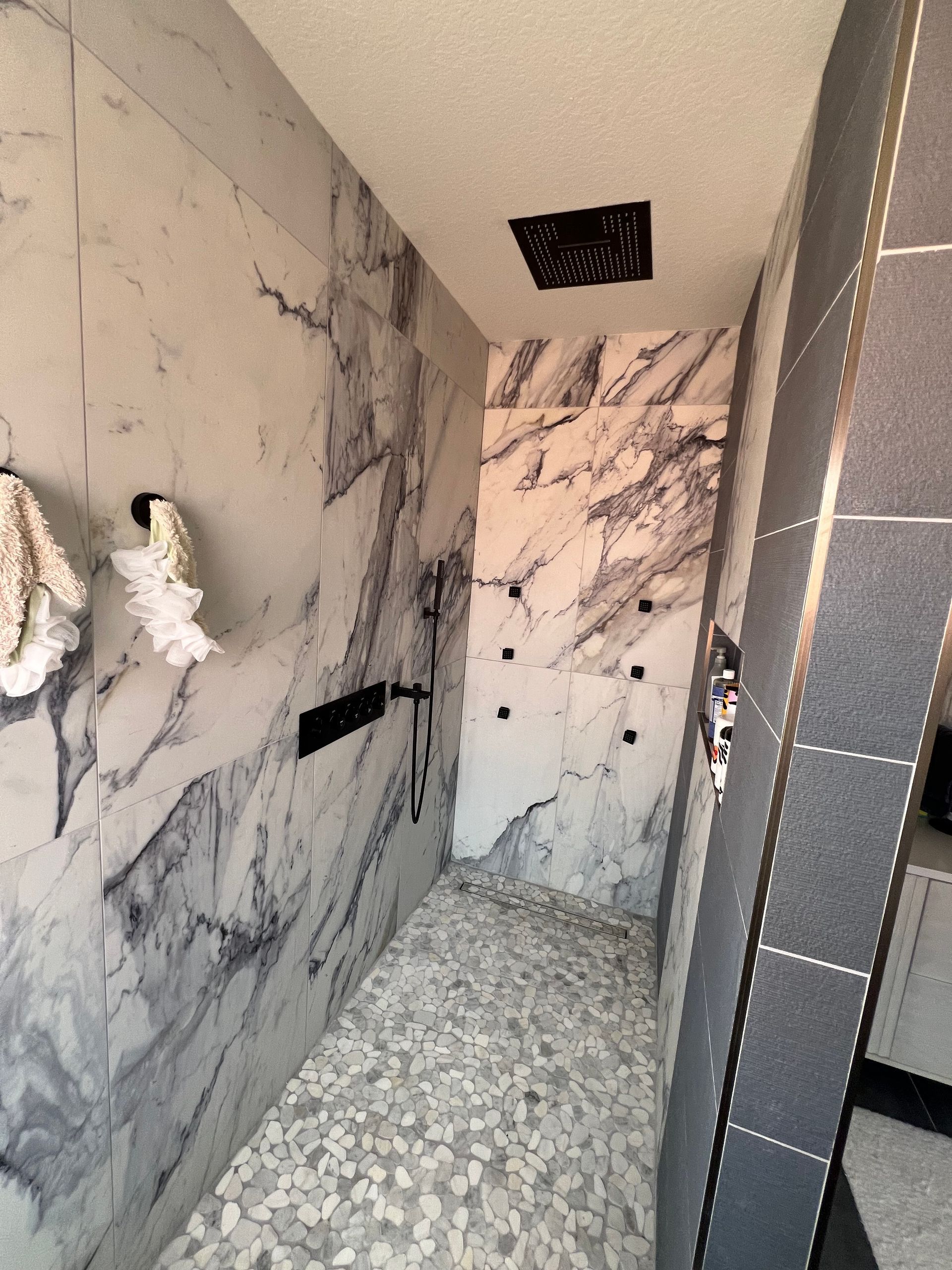 Marble shower with pebble floor, black fixtures, and a dark ceiling vent.