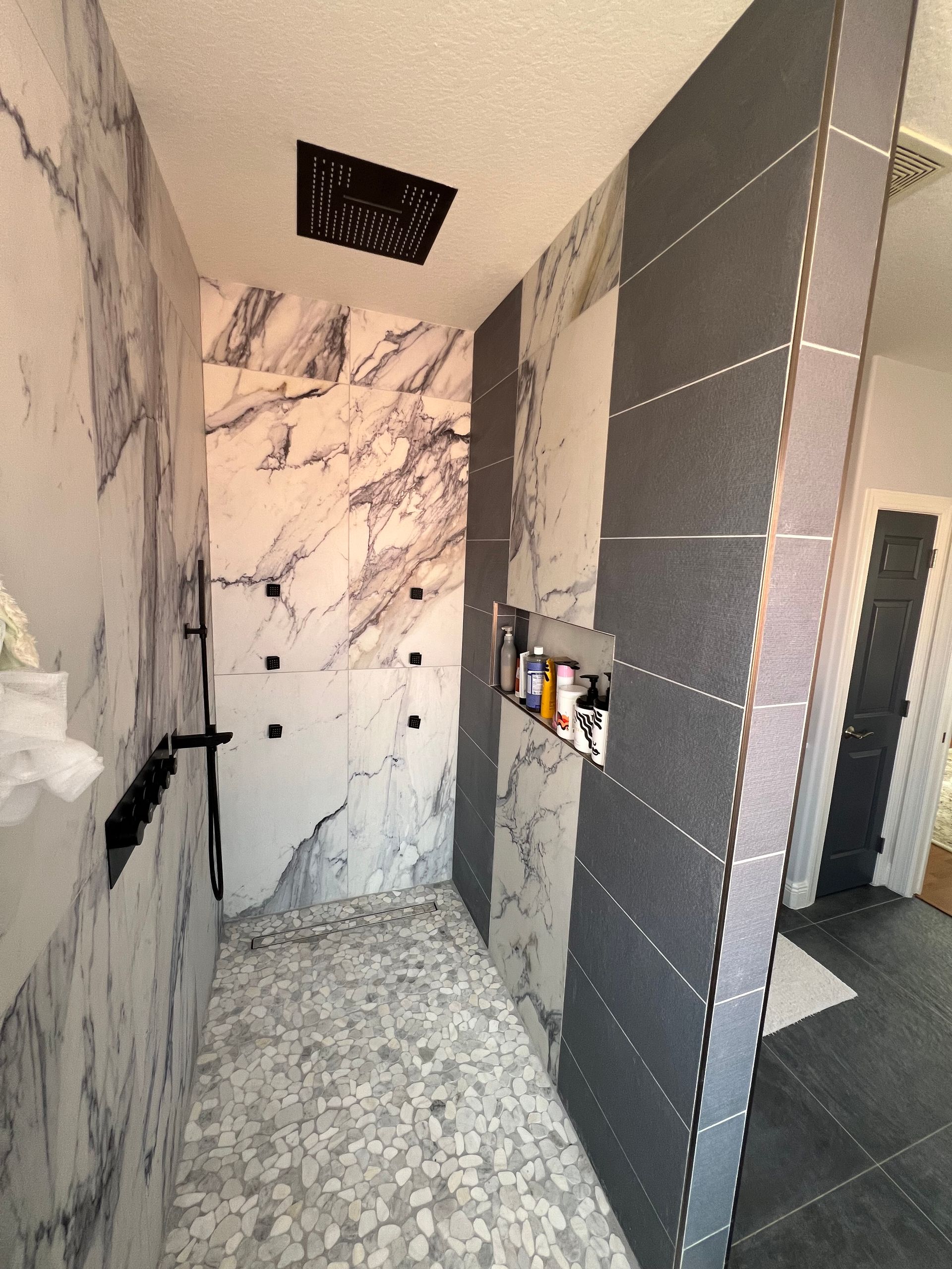 A modern shower with marble and gray tile walls, black fixtures, and pebble tile flooring.