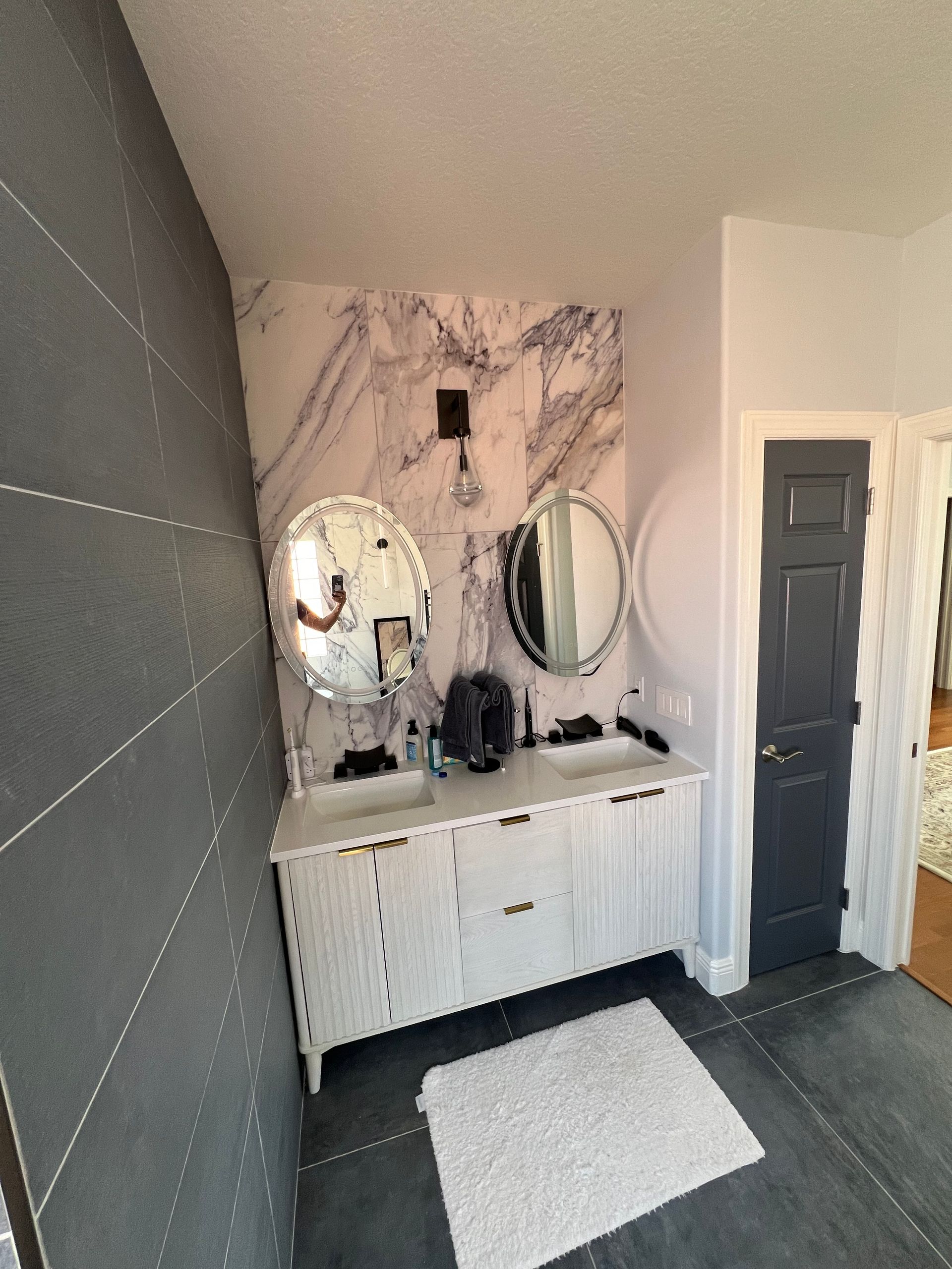 Modern bathroom: white vanity with two mirrors, marble accent wall, gray tiled floor, dark gray door.