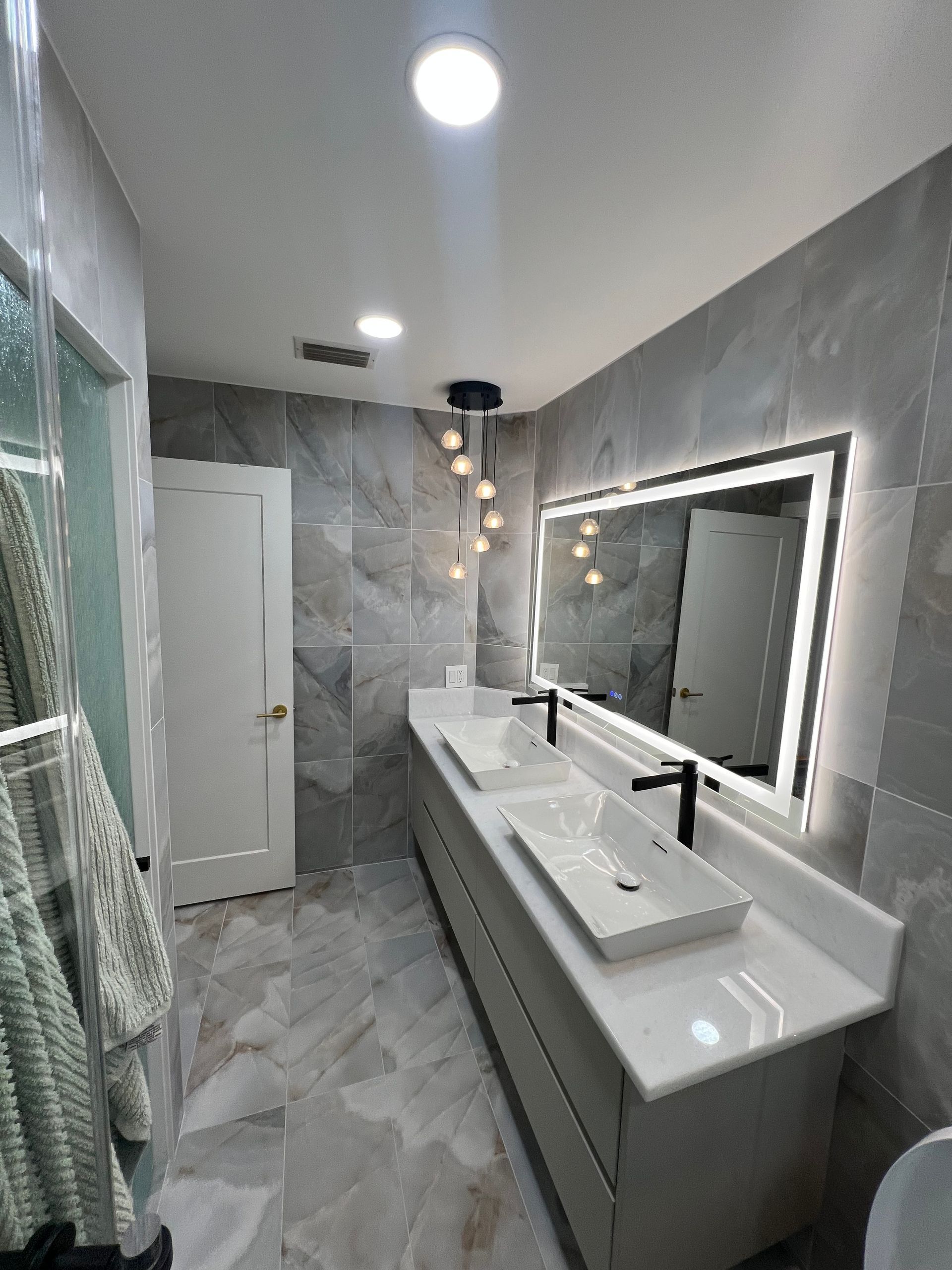 Modern bathroom with double sinks, illuminated mirror, light gray tiles, and white door.