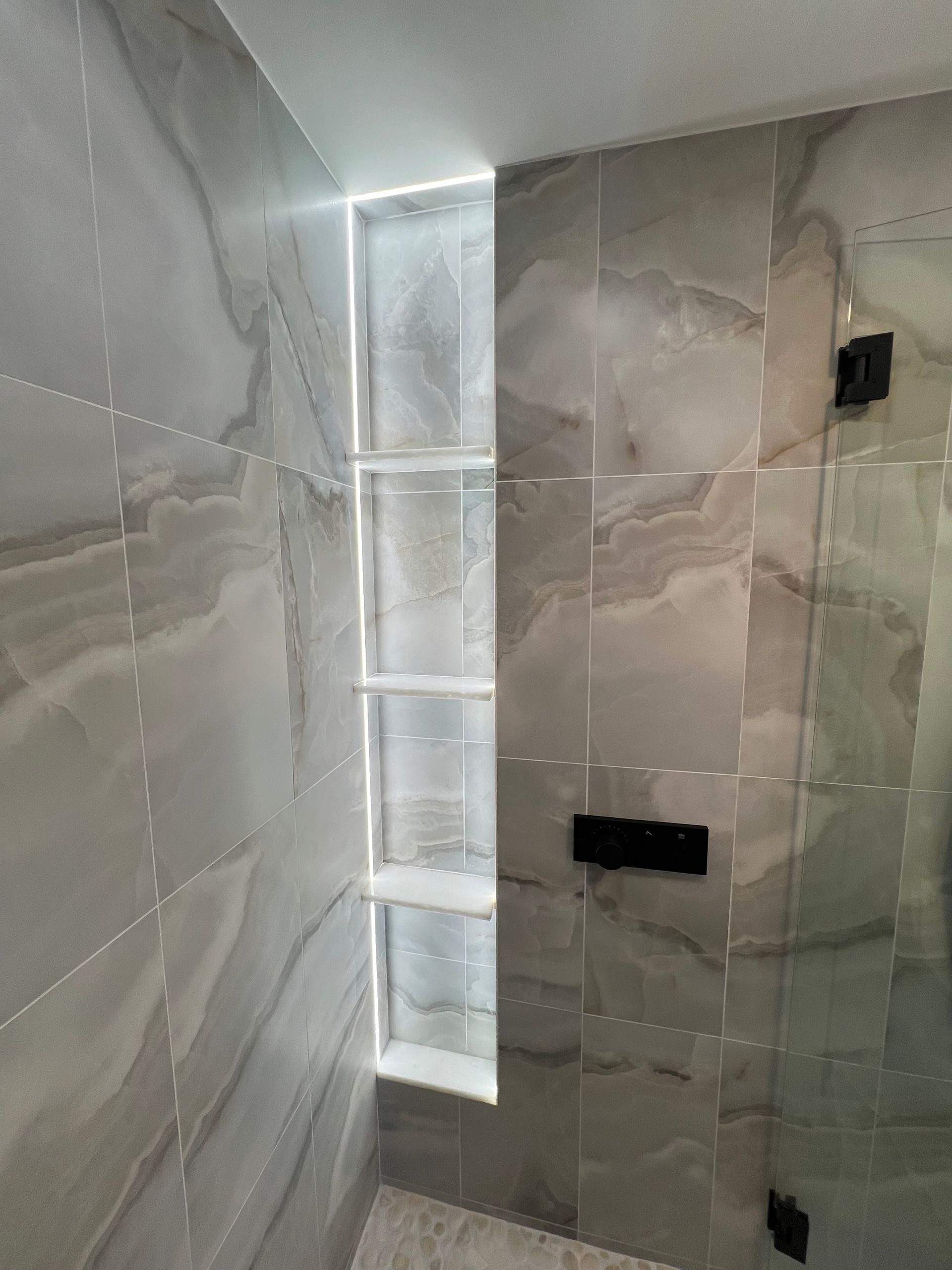 A shower interior with built-in shelves and gray marble-like tiles, illuminated by a strip light.