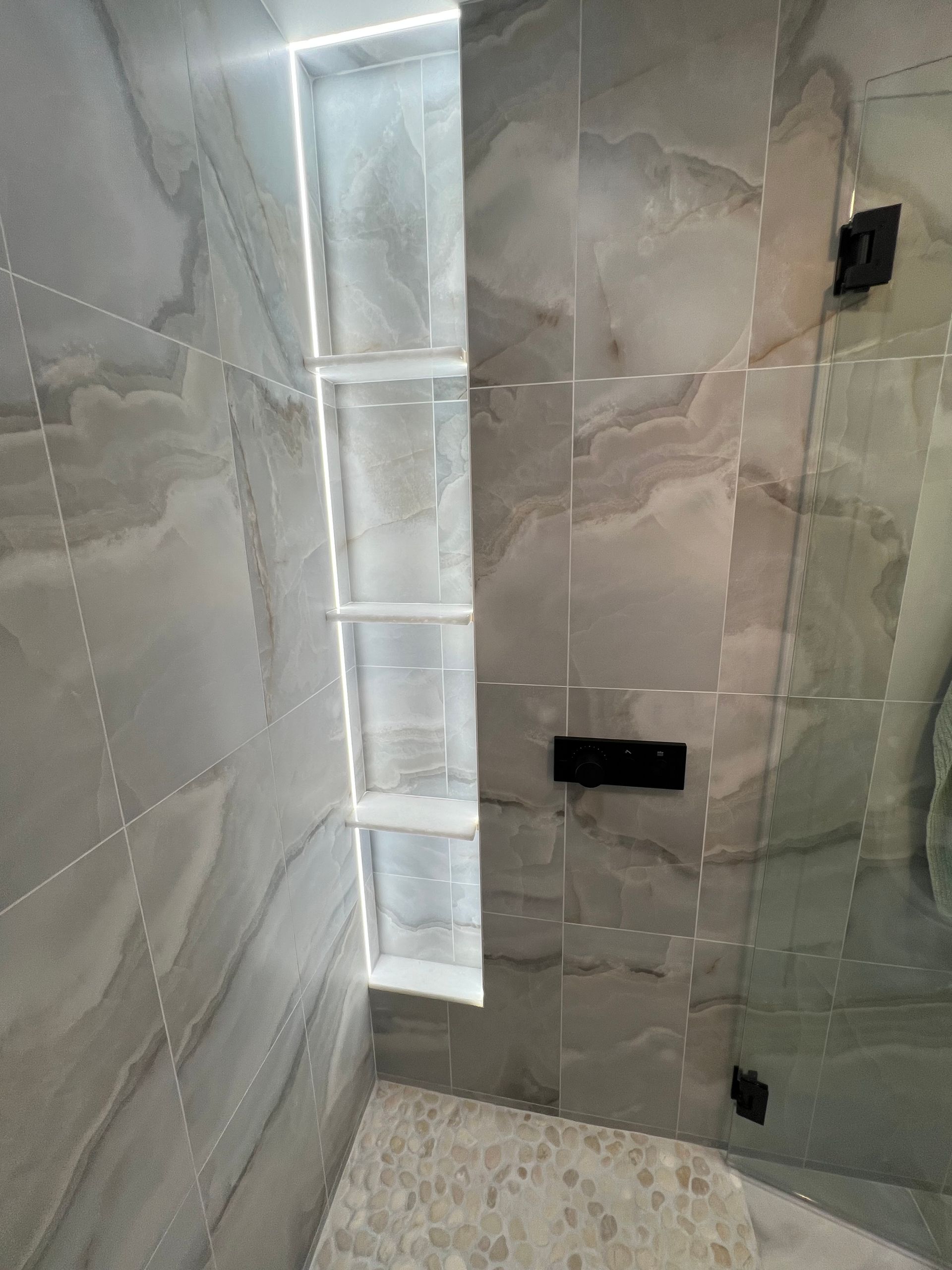 Tiled shower interior with recessed, lit shelves. Gray marble walls, glass shower door, and a pebble floor.